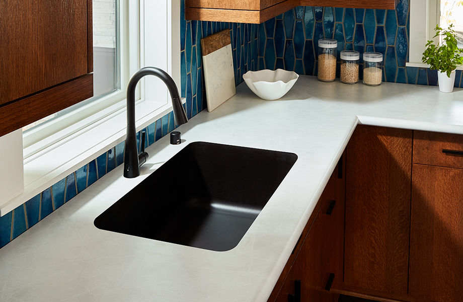 Kitchen with brown cabinets, blue tile backsplash, White Alabaster 180fx Laminate countertops, black sink and faucet, and jars of grains